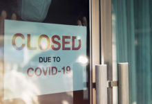 Business office or store shop is closed/bankrupt business due to the effect of novel Coronavirus (COVID-19) pandemic. Unidentified person wearing mask hanging closed sign in background on front door.