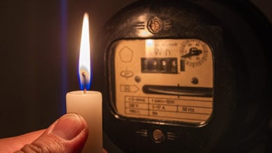 Power outage or blackout concept image. Person's hand with candle shining light in the dark near electricity meter at home.