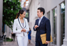 Portrait of two young, confident and attractive Asian business people standing in a city during the day. They are both professionally dressed in suits and are smiling as they talk to one another. ; Shutterstock ID 1450298177