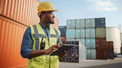 Smiling Portrait of a Handsome African American Black Industrial Engineer in Yellow Hard Hat and Safety Vest Working on Tablet Computer. Foreman or Supervisor in Container Terminal.