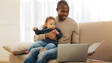 Paternal leave. Cheerful dark-eyed bearded afro-american man smiling and holding his son on his lap while working on the laptop and holding a sheet of paper