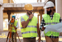 Three men in hard hats on a building site, one holding plans