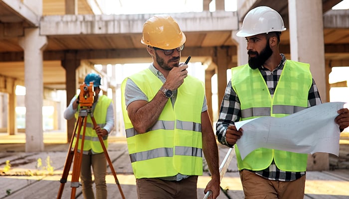 Three men in hard hats on a building site, one holding plans